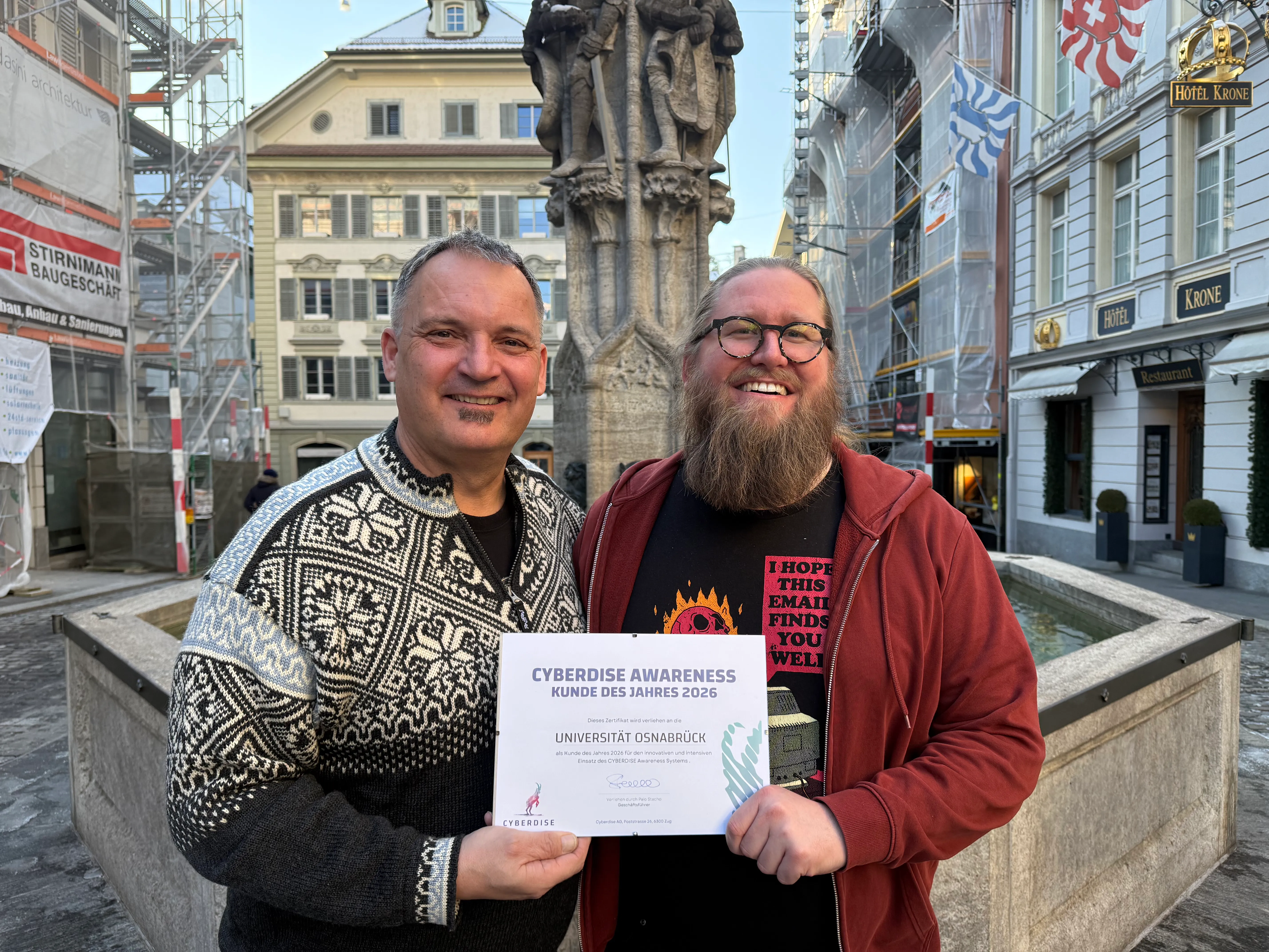 Two men at an old-town fountain holding the Awareness Customer of the Year 2026 certificate for Universität Osnabrück